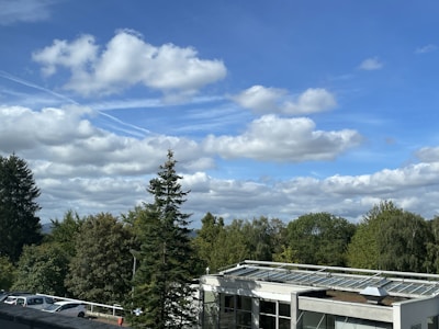 A landscape featuring a clear blue sky with scattered fluffy white clouds. In the foreground, a mix of evergreen and deciduous trees is visible. Below, there is a building with a modern design, featuring a flat roof with glass panels. Several cars are parked nearby, hinting at a parking area.