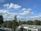 A landscape featuring a clear blue sky with scattered fluffy white clouds. In the foreground, a mix of evergreen and deciduous trees is visible. Below, there is a building with a modern design, featuring a flat roof with glass panels. Several cars are parked nearby, hinting at a parking area.