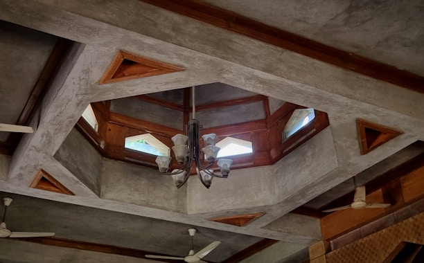A geometric ceiling design features triangular and polygonal shapes with wooden accents. The ceiling includes several glass panels that allow natural light to filter through. A chandelier with frosted glass shades hangs at the center, surrounded by ceiling fans.