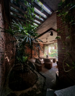 A rustic interior space featuring exposed brick walls and a natural skylight with hanging green plants. There's a round wooden table surrounded by stools in the center, creating a cozy dining or meeting area. The natural lighting and abundant greenery contribute to a harmonious and tranquil ambiance.