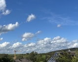 A wide landscape with a clear blue sky filled with scattered fluffy clouds. Below, a green expanse of fields and trees is visible. A construction crane and solar panels on a rooftop can be seen, suggesting a mix of nature and technology.