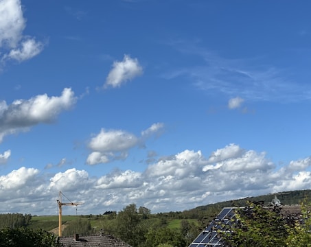 A wide landscape with a clear blue sky filled with scattered fluffy clouds. Below, a green expanse of fields and trees is visible. A construction crane and solar panels on a rooftop can be seen, suggesting a mix of nature and technology.
