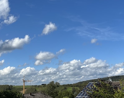A wide landscape with a clear blue sky filled with scattered fluffy clouds. Below, a green expanse of fields and trees is visible. A construction crane and solar panels on a rooftop can be seen, suggesting a mix of nature and technology.