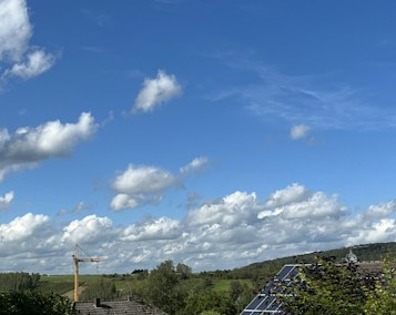 A wide landscape with a clear blue sky filled with scattered fluffy clouds. Below, a green expanse of fields and trees is visible. A construction crane and solar panels on a rooftop can be seen, suggesting a mix of nature and technology.