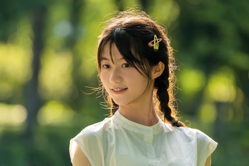 A young woman with braided hair and a hairpin is smiling softly. She is wearing a sleeveless light-colored top. The background is blurred greenery, suggesting a natural or outdoor setting.