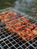 Grilled skewers of marinated meat are cooking on a metal barbecue grill, with visible grill marks and charred edges. Smoke drifts upwards, adding a rustic outdoor cooking atmosphere. The meat is evenly spaced and appears to be well-seasoned.