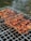 Grilled skewers of marinated meat are cooking on a metal barbecue grill, with visible grill marks and charred edges. Smoke drifts upwards, adding a rustic outdoor cooking atmosphere. The meat is evenly spaced and appears to be well-seasoned.