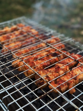 Grilled skewers of marinated meat are cooking on a metal barbecue grill, with visible grill marks and charred edges. Smoke drifts upwards, adding a rustic outdoor cooking atmosphere. The meat is evenly spaced and appears to be well-seasoned.