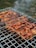 Grilled skewers of marinated meat are cooking on a metal barbecue grill, with visible grill marks and charred edges. Smoke drifts upwards, adding a rustic outdoor cooking atmosphere. The meat is evenly spaced and appears to be well-seasoned.