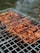 Grilled skewers of marinated meat are cooking on a metal barbecue grill, with visible grill marks and charred edges. Smoke drifts upwards, adding a rustic outdoor cooking atmosphere. The meat is evenly spaced and appears to be well-seasoned.