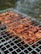 Grilled skewers of marinated meat are cooking on a metal barbecue grill, with visible grill marks and charred edges. Smoke drifts upwards, adding a rustic outdoor cooking atmosphere. The meat is evenly spaced and appears to be well-seasoned.