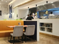 A modern restaurant interior featuring a table with two grey chairs on a wooden floor. The table is set with glass bottles and napkins. Behind the seating area is an open kitchen with chefs in white uniforms and tall hats. The kitchen has metallic surfaces and hanging lights. The wall has decorative patterns and artwork, creating an elegant setting.