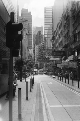 A busy urban street scene with high-rise buildings and bustling pedestrian activity. The street is lined with tall buildings featuring multiple signs and advertisements in different languages, suggesting a vibrant commercial district. Pedestrians walk along the sidewalks, some carrying bags and others pausing at intersections. The presence of a traffic light and several vehicles, including a bus, indicates a well-traveled city area.