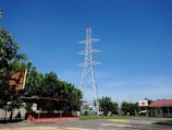 A towering metal electricity pylon stands against a clear blue sky, surrounded by green trees. A basketball court with hoops is in the foreground, and there are small buildings with red roofs in the background. Power lines extend from the pylon across the image.