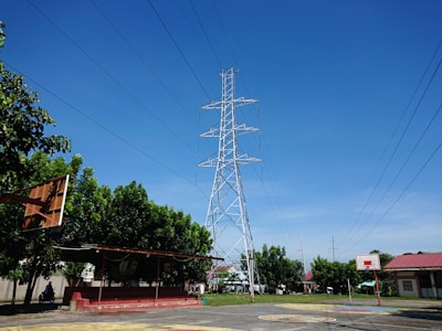 A towering metal electricity pylon stands against a clear blue sky, surrounded by green trees. A basketball court with hoops is in the foreground, and there are small buildings with red roofs in the background. Power lines extend from the pylon across the image.