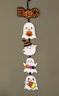 A vertical decoration featuring four cute, cartoonish ghosts with varying expressions and accessories. At the top, an orange and black glittery 'BOO' sign is displayed. Each ghost holds Halloween-themed items such as a pumpkin, a witch hat, candy letters spelling 'BOO', and candy corn.