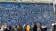 A large blue wall covered in various languages expressing the phrase 'I love you' in different scripts. The wall is sectioned into multiple tiles with white and red writings, each tile featuring the phrase in a unique language. At the base of the wall, a group of people is gathered, some interacting with each other, others taking photos.