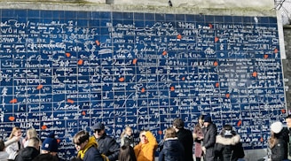 A large blue wall covered in various languages expressing the phrase 'I love you' in different scripts. The wall is sectioned into multiple tiles with white and red writings, each tile featuring the phrase in a unique language. At the base of the wall, a group of people is gathered, some interacting with each other, others taking photos.
