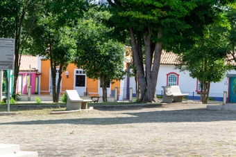 A peaceful urban park scene featuring colorful buildings in the background, including pink, orange, and white facades with tiled roofs. Trees provide shade and green foliage, while a few concrete benches are arranged on a stone-floored area. The image evokes a sense of calm and community.
