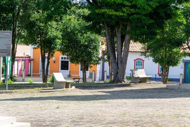 A peaceful urban park scene featuring colorful buildings in the background, including pink, orange, and white facades with tiled roofs. Trees provide shade and green foliage, while a few concrete benches are arranged on a stone-floored area. The image evokes a sense of calm and community.