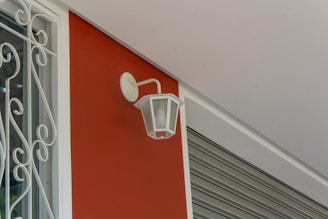 A white, vintage-style wall lantern is mounted on a smooth, red wall. To the left, there is a window with decorative white wrought iron bars. The ceiling is light grey, and a metallic roller shutter is partially visible on the right side.