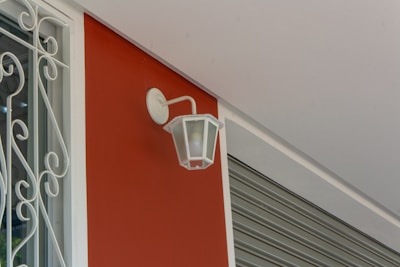 A white, vintage-style wall lantern is mounted on a smooth, red wall. To the left, there is a window with decorative white wrought iron bars. The ceiling is light grey, and a metallic roller shutter is partially visible on the right side.