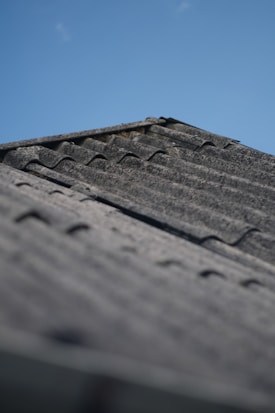 A close-up view of a grey corrugated roof under a clear blue sky. The surface of the roof shows signs of wear and age, with slight discoloration and moss growth along the edges.