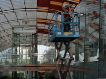 A worker uses a mobile platform lift to clean or repair the glass facade of a modern building with a curved, partially transparent roof and a background of urban structures.