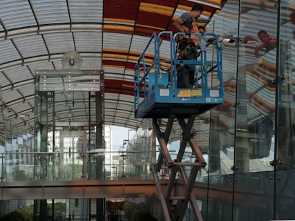 A worker uses a mobile platform lift to clean or repair the glass facade of a modern building with a curved, partially transparent roof and a background of urban structures.