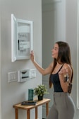 A woman stands in an interior setting, interacting with a white board mounted on the wall. She is holding a set of keys in one hand and appears to be engaged with a list of errands written on the board. Below the board, there is a small wooden table with a plant and a green candle resting on it. Light switches are visible on the wall.