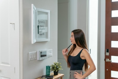 A woman with long brown hair is looking at a list written on a whiteboard mounted on the wall. She is holding a marker near her face, appearing thoughtful. The board lists errands like grocery and yoga. Below the board are a thermostat and a small shelf with a plant and a container.