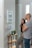 A man and a woman stand together looking at a whiteboard mounted on a wall, which lists errands such as grocery and yoga at 6pm. The interior of the room is modern, with a small wooden table that holds a plant, a green candle, and some keys. The wall features light switches and an electronic display panel.