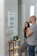 A man and a woman stand together looking at a whiteboard mounted on a wall, which lists errands such as grocery and yoga at 6pm. The interior of the room is modern, with a small wooden table that holds a plant, a green candle, and some keys. The wall features light switches and an electronic display panel.