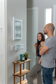 A man and a woman stand together looking at a whiteboard mounted on a wall, which lists errands such as grocery and yoga at 6pm. The interior of the room is modern, with a small wooden table that holds a plant, a green candle, and some keys. The wall features light switches and an electronic display panel.