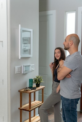 A man and a woman stand together looking at a whiteboard mounted on a wall, which lists errands such as grocery and yoga at 6pm. The interior of the room is modern, with a small wooden table that holds a plant, a green candle, and some keys. The wall features light switches and an electronic display panel.