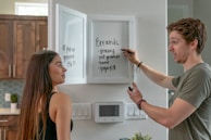 A man and a woman are in a modern kitchen. The man is writing on the inner side of a white cabinet door with a marker, listing errands like grocery shopping and yoga. The woman is standing beside him, looking at the list. The kitchen features wooden cabinets, white walls, an electronic device on the wall, and some greenery.