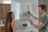 A man and a woman are in a modern kitchen. The man is writing on the inner side of a white cabinet door with a marker, listing errands like grocery shopping and yoga. The woman is standing beside him, looking at the list. The kitchen features wooden cabinets, white walls, an electronic device on the wall, and some greenery.