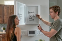 A man and a woman are in a modern kitchen. The man is writing on the inner side of a white cabinet door with a marker, listing errands like grocery shopping and yoga. The woman is standing beside him, looking at the list. The kitchen features wooden cabinets, white walls, an electronic device on the wall, and some greenery.