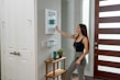 A woman with long hair, dressed in a black tank top and gray pants, is interacting with a smart home device mounted on a wall. She is holding a tablet in her left hand. The setting appears to be a modern home entryway with a wooden door featuring horizontal glass panels. A small side table near the wall supports a plant and some decorative items.