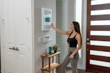 A woman with long hair, dressed in a black tank top and gray pants, is interacting with a smart home device mounted on a wall. She is holding a tablet in her left hand. The setting appears to be a modern home entryway with a wooden door featuring horizontal glass panels. A small side table near the wall supports a plant and some decorative items.