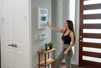 A woman with long hair, dressed in a black tank top and gray pants, is interacting with a smart home device mounted on a wall. She is holding a tablet in her left hand. The setting appears to be a modern home entryway with a wooden door featuring horizontal glass panels. A small side table near the wall supports a plant and some decorative items.