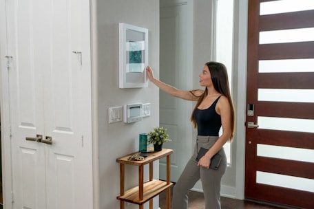 A woman with long hair, dressed in a black tank top and gray pants, is interacting with a smart home device mounted on a wall. She is holding a tablet in her left hand. The setting appears to be a modern home entryway with a wooden door featuring horizontal glass panels. A small side table near the wall supports a plant and some decorative items.