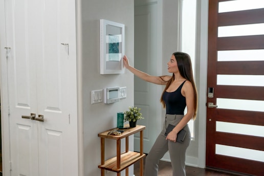 A woman with long hair, dressed in a black tank top and gray pants, is interacting with a smart home device mounted on a wall. She is holding a tablet in her left hand. The setting appears to be a modern home entryway with a wooden door featuring horizontal glass panels. A small side table near the wall supports a plant and some decorative items.