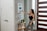 A woman with long hair, dressed in a black tank top and gray pants, is interacting with a smart home device mounted on a wall. She is holding a tablet in her left hand. The setting appears to be a modern home entryway with a wooden door featuring horizontal glass panels. A small side table near the wall supports a plant and some decorative items.