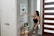 A woman with long hair, dressed in a black tank top and gray pants, is interacting with a smart home device mounted on a wall. She is holding a tablet in her left hand. The setting appears to be a modern home entryway with a wooden door featuring horizontal glass panels. A small side table near the wall supports a plant and some decorative items.