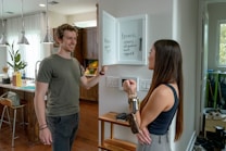 Two people are interacting in a modern kitchen. A woman is holding a set of keys and facing a man, who appears to be smiling. There is a whiteboard on the wall listing errands such as groceries and yoga. The kitchen is well-lit with large windows and pendant lights hanging over the counter.