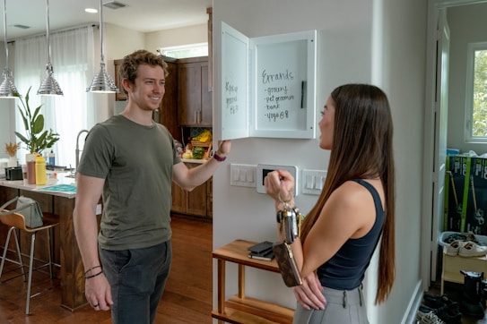Two people are interacting in a modern kitchen. A woman is holding a set of keys and facing a man, who appears to be smiling. There is a whiteboard on the wall listing errands such as groceries and yoga. The kitchen is well-lit with large windows and pendant lights hanging over the counter.