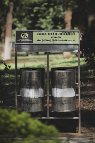 Two metallic bins positioned side by side in an outdoor setting with green foliage in the background. The bins have a perforated design, surrounded by a supporting metal frame with a sign in Hindi above them.