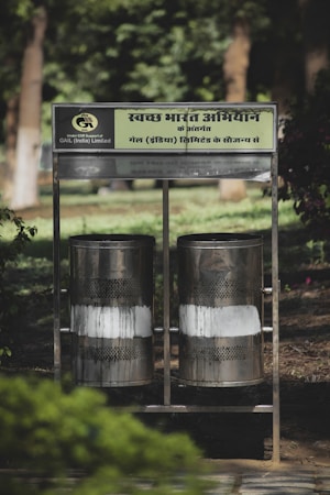 Two metallic bins positioned side by side in an outdoor setting with green foliage in the background. The bins have a perforated design, surrounded by a supporting metal frame with a sign in Hindi above them.