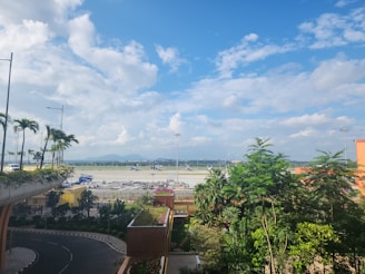 A corner NA plot with a paved road access and nearby greenery under a clear sunny sky.